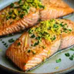 Air fryer salmon lemon - the image shows two pieces of salmon on a white plate. the salmon is cooked to a golden brown color and is garnished with chopped herbs and lemon zest. the plate is sitting on a dark grey countertop. the background is blurred, making the salmon the focal point of the image.