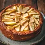 Apple cake korean - the image is of a round cake on a blue plate. the cake appears to be a tart with a golden brown crust and a layer of caramelized apples on top. the apples are arranged in a circular pattern on the top of the cake, with some overlapping each other. the plate is placed on a wooden table with a rustic background. the overall mood of the image is warm and inviting.