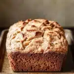 Apple cinnamon muffin loaf - the image is a close-up of a freshly baked loaf of bread on a metal tray. the bread appears to be golden brown in color and has a crumbly texture on top. the tray is placed on a wooden table with a rustic background. the loaf is slightly tilted to the side, and the edges of the bread are slightly curled. the overall mood of the image is warm and inviting.