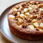 Apple mug cake - the image is a close-up of a round cake on a white plate. the cake appears to be freshly baked and has a golden brown crust. on top of the cake, there are small pieces of sliced apples arranged in a criss-cross pattern. the apples are cut into small cubes and are scattered throughout the cake. the plate is sitting on a wooden table with a light-colored surface. the background is blurred, making the cake the focal point of the image.