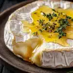 Baked brie with honey and thyme - the image is a close-up of a dessert on a brown plate. the dessert appears to be a brie, a type of camembert cheese, with a golden-brown crust and a creamy yellow filling. the filling is drizzled over the top, creating a smooth and creamy texture. on top of the filling, there is a sprig of fresh thyme, adding a pop of green color to the dish. the plate is sitting on a wooden table with a white cloth napkin in the background.