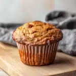 Banana bread kodiak muffins - the image shows a freshly baked muffin on a wooden cutting board. the muffin is golden brown in color and has a crumbly texture. it is sitting on a gray cloth napkin on the right side of the cutting board, and the background is blurred, making the muffin the focal point of the image.