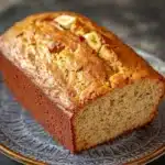 Banana cake loaf - the image shows a freshly baked loaf of banana bread on a blue and white patterned plate. the bread is golden brown in color and has a slightly crisp crust on top. it is topped with sliced bananas, giving it a crunchy texture. the plate is sitting on a dark grey countertop. the background is blurred, making the bread the focal point of the image.