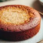 Banana cake no butter - the image is a close-up of a round cake on a white plate. the cake appears to be a chocolate cake with a golden brown crust on top. the top of the cake is covered in a light dusting of powdered sugar, giving it a shiny appearance. the plate is sitting on a wooden table with a blurred background of greenery. a fork is resting on the plate next to the cake.