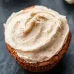 Banana muffin frosting - the image shows a close-up of a cupcake with a generous layer of frosting on top. the frosting appears to be a light beige color and has a smooth texture. the cupcake is sitting on a dark grey textured surface, possibly a table or countertop. in the background, there are two whole bananas, one on the right side of the image and the other on the left side. the background is blurred, but it seems to be an indoor setting.