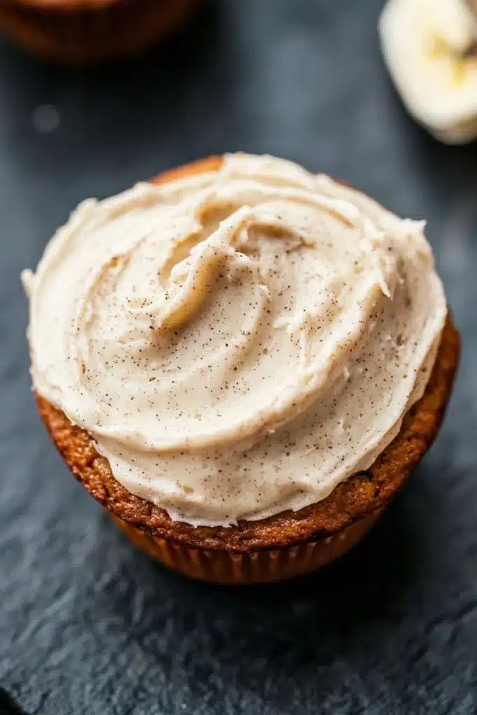Banana muffin frosting - the image shows a close-up of a cupcake with a generous layer of frosting on top. the frosting appears to be a light beige color and has a smooth texture. the cupcake is sitting on a dark grey textured surface, possibly a table or countertop. in the background, there are two whole bananas, one on the right side of the image and the other on the left side. the background is blurred, but it seems to be an indoor setting.