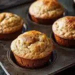 Banana muffin sour cream - the image shows a muffin tray with six freshly baked muffins in it. the muffins are golden brown in color and have a crumbly texture. they are arranged in a single layer in the tray, with the topmost muffin slightly overlapping the bottom one. the tray is placed on a wooden table, and the background is blurred, making the muffins the focal point of the image.