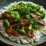 Beef and broccoli stir fry rice - the image shows a plate of stir-fried beef and broccoli on a bed of white rice. the beef is cooked medium-rare and appears to be seasoned with herbs and spices. the broccoli is bright green and looks fresh and healthy. the rice is fluffy and white, and the dish is served on a dark blue plate with a brown rim. the background is blurred, making the plate the focal point of the image.