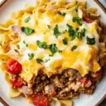 Beef casserole with ground beef and noodles - the image shows a plate of pasta dish with a layer of ground beef and cheese on top. the dish is served on a white marble countertop with a gray and white striped napkin on the side. the pasta appears to be fettuccine alfredo, which is a type of italian pasta. the top of the dish is covered in melted cheese and garnished with chopped parsley. there are also chunks of red bell peppers scattered throughout the dish. the overall color scheme of the image is warm and inviting.