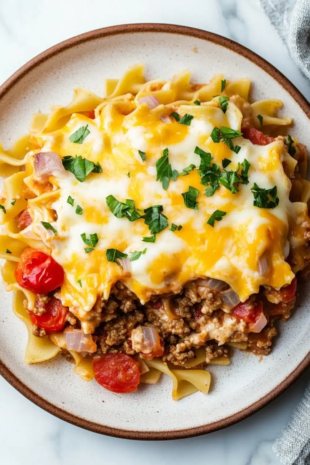 Beef casserole with ground beef and noodles - the image shows a plate of pasta dish with a layer of ground beef and cheese on top. the dish is served on a white marble countertop with a gray and white striped napkin on the side. the pasta appears to be fettuccine alfredo, which is a type of italian pasta. the top of the dish is covered in melted cheese and garnished with chopped parsley. there are also chunks of red bell peppers scattered throughout the dish. the overall color scheme of the image is warm and inviting.