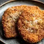 Beef chicken cutlets - the image shows two pieces of breaded fish fillets on a black plate. the fillets are golden brown and appear to be seasoned with herbs and spices. the plate is round and has a textured surface. the background is blurred, but it appears to be a dark countertop. the overall mood of the image is rustic and appetizing.