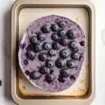 Blueberry muffin smoothie - the image shows a rectangular baking tray with a freshly baked blueberry cake on it. the cake is a light brown color and has a smooth texture. on top of the cake, there are a few fresh blueberries scattered around the tray. the blueberries are a deep purple color and appear to be ripe and juicy. the tray is sitting on a white marble countertop.