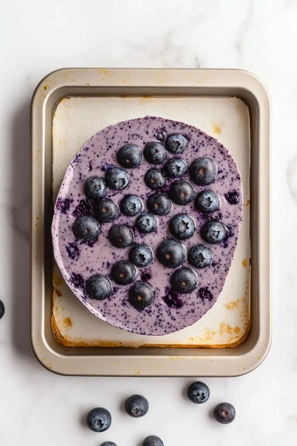Blueberry muffin smoothie - the image shows a rectangular baking tray with a freshly baked blueberry cake on it. the cake is a light brown color and has a smooth texture. on top of the cake, there are a few fresh blueberries scattered around the tray. the blueberries are a deep purple color and appear to be ripe and juicy. the tray is sitting on a white marble countertop.