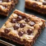 Chewy chocolate chip cookie bars - the image shows four square-shaped chocolate chip cookie bars on a black slate tray. the bars are golden brown in color and have a crumbly texture. they are topped with chocolate chips and drizzled with a dark chocolate sauce. the tray is lined with parchment paper and there are a few more bars visible in the background. the overall aesthetic of the image is rustic and appetizing.
