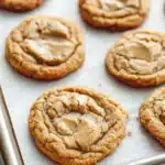 Chewy peanut butter cookie - the image shows a tray of freshly baked peanut butter cookies. the cookies are round and golden brown in color, with a crumbly texture on top. they are arranged in a single layer on the tray, with some overlapping each other. the tray is lined with parchment paper, and the cookies appear to be freshly baked. the background is blurred, but it appears to be a kitchen countertop.