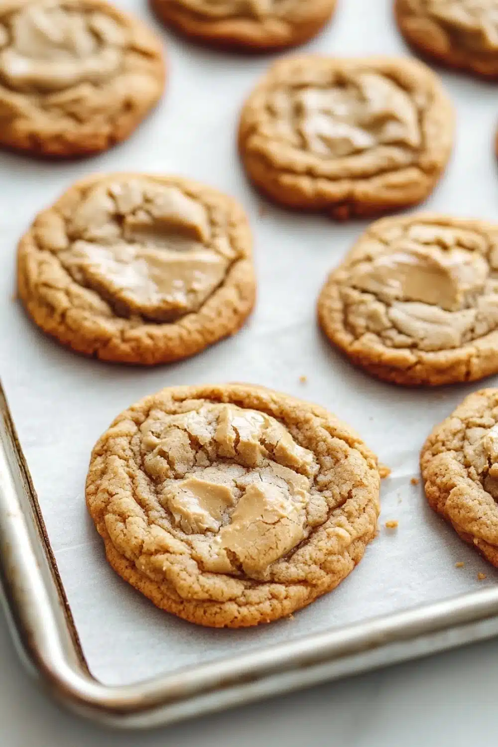 Chewy peanut butter cookie - the image shows a tray of freshly baked peanut butter cookies. the cookies are round and golden brown in color, with a crumbly texture on top. they are arranged in a single layer on the tray, with some overlapping each other. the tray is lined with parchment paper, and the cookies appear to be freshly baked. the background is blurred, but it appears to be a kitchen countertop.