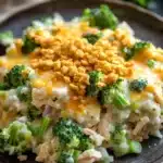 Chicken broccoli rice casserole trisha yearwood - the image shows a plate of food with a serving of rice and broccoli. the rice appears to be cooked and has a golden brown color. the broccoli is bright green and looks fresh. on top of the rice, there is a layer of melted cheese and crumbled breadcrumbs. the dish is garnished with a sprig of parsley. the plate is brown and the background is blurred, but it seems to be a wooden table.