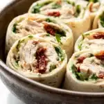 Chicken caesar salad pinwheels - the image shows a bowl filled with rolled up wraps. the wraps are golden brown and appear to be filled with a mixture of chicken, herbs, and sun-dried tomatoes. the chicken is cut into thin strips and is arranged in a circular pattern. the herbs and tomatoes are scattered throughout the wraps, adding a pop of green color to the dish. the bowl is made of ceramic and has a textured surface. the background is blurred, but it appears to be a white countertop.