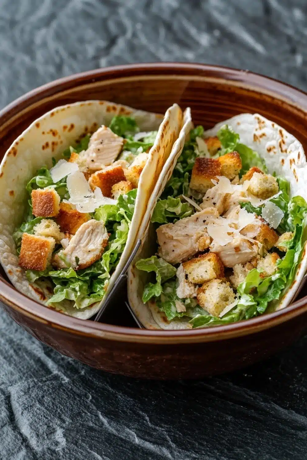 Chicken caesar salad tacos - the image shows two tacos in a brown ceramic bowl. the tacos are filled with a salad of green lettuce, croutons, and shredded chicken. the chicken appears to be seasoned with herbs and spices, and there is a sprinkle of parmesan cheese on top. the bowl is sitting on a dark grey countertop.