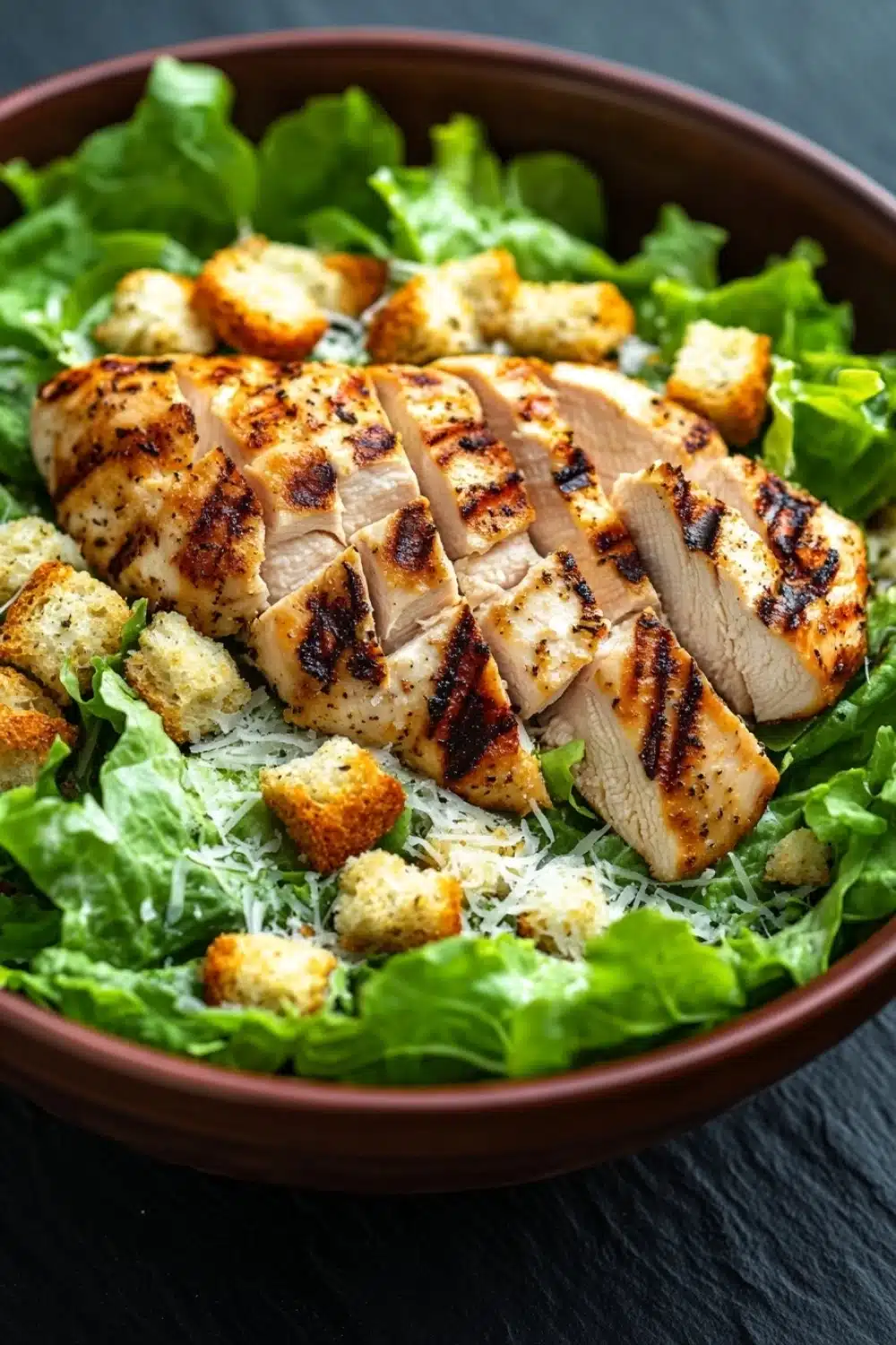 Chicken caesar salad - the image is a close-up of a salad in a brown bowl. the salad is made up of lettuce leaves and croutons, and there are several pieces of grilled chicken arranged in a circular pattern on top. the chicken appears to be seasoned with herbs and spices, and it is drizzled with a light dressing. the bowl is sitting on a dark grey countertop.