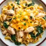 Chicken noodle skillet with sauce - the image is a close-up of a bowl of pasta dish. the pasta appears to be fettuccine alfredo, with chunks of chicken and spinach mixed in. the dish is garnished with a dollop of melted cheese and a sprinkle of fresh parsley. the bowl is white with a brown rim and is sitting on a white marble countertop. there are a few sprigs of parsley scattered around the bowl.