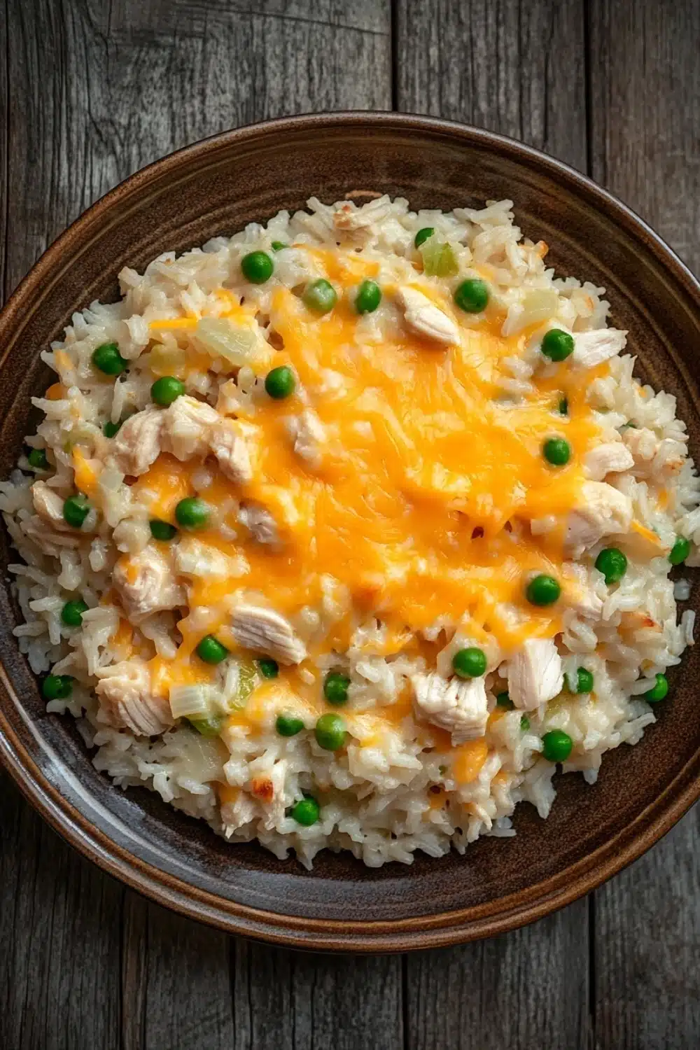 Chicken rice casserole knorr - the image is a close-up of a bowl of rice dish. the rice appears to be white and fluffy, and it is topped with chunks of chicken and green peas. the dish is served in a brown ceramic bowl with a dark brown rim. the background is a wooden table with a rustic texture. the overall color scheme of the dish is warm and inviting.