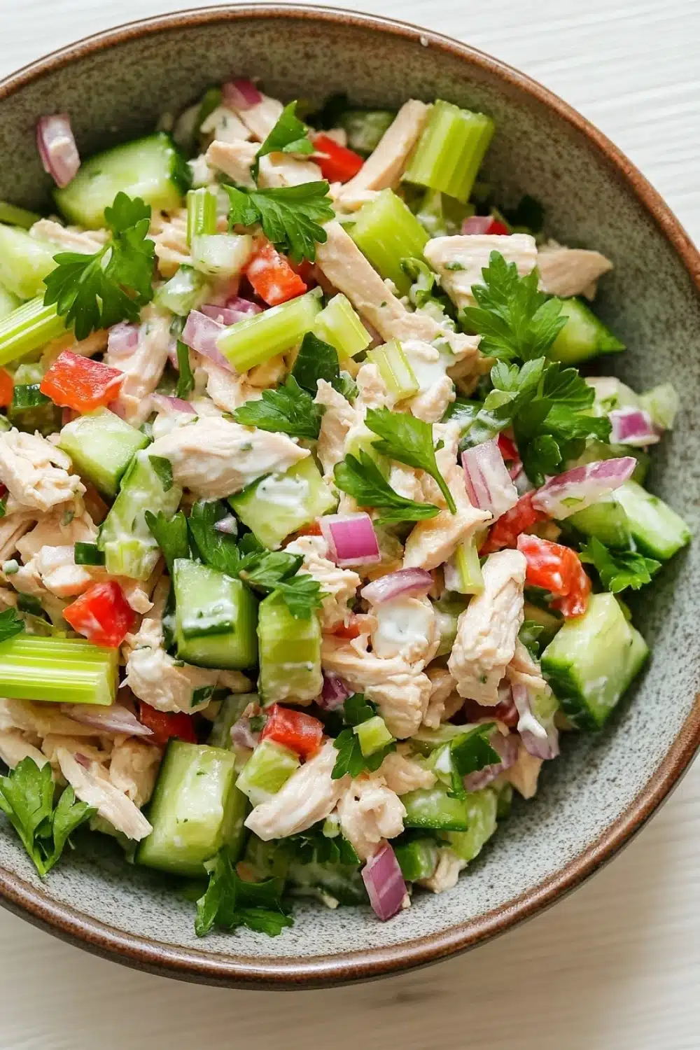 Chicken salad finely chopped - the image is a close-up of a bowl of salad. the bowl is made of ceramic and has a light brown color. the salad is made up of diced celery, red onions, and chunks of chicken. the chicken is mixed with the vegetables, creating a colorful and appetizing appearance. there are also some fresh cilantro leaves scattered throughout the salad, adding a pop of green color to the dish.
