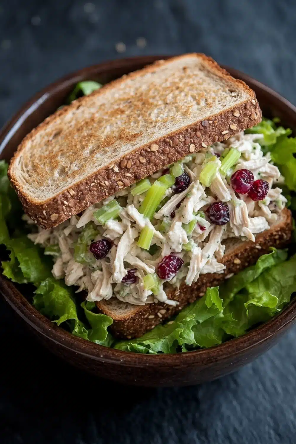 Chicken salad sandwich with cranberries - the image shows a wooden bowl with a sandwich in it. the sandwich is made with two slices of toasted bread and is filled with a salad. the salad appears to be made with chicken, celery, and cranberries. the bowl is garnished with fresh green lettuce leaves. the background is a dark grey countertop.