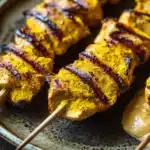 Chicken satay skewers appetizers - the image shows a plate of grilled chicken skewers on a wooden table. the skewers are arranged in a row and are covered in a golden-brown sauce. the sauce appears to be a combination of orange and yellow colors, possibly from the spices used in the dish. the plate is brown and has a speckled texture. the background is blurred, but it seems to be an indoor setting with a wooden surface.