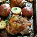 Chicken sheet pan dinner potatoes - the image shows a baking tray filled with roasted chicken and potatoes. the chicken is golden brown and appears to be seasoned with herbs and spices. the potatoes are red and have a crispy texture. there are also some garlic cloves scattered around the tray. the tray is lined with parchment paper and is sitting on a wooden table. the overall appearance of the dish is savory and appetizing.