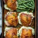Chicken thigh dinner and sides - the image shows a rectangular baking tray with four pieces of chicken thighs on top of a bed of mashed potatoes. the chicken thighs are golden brown and appear to be seasoned with herbs and spices. the mashed potatoes are white and fluffy, and there are a few green beans scattered around the tray. the tray is placed on a wooden table with a blue napkin on the side. the overall presentation of the dish is elegant and appetizing.