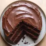 Chocolate cake normal design - the image is of a round chocolate cake on a white plate. the cake has a thick layer of chocolate frosting on top, with a swirl pattern on the sides. the frosting appears to be smooth and glossy, and the cake is arranged in a neat and orderly manner. there is a slice taken out of the cake, revealing the layers of chocolate cake. the plate is sitting on a wooden table, and there are a few crumbs scattered around the plate.
