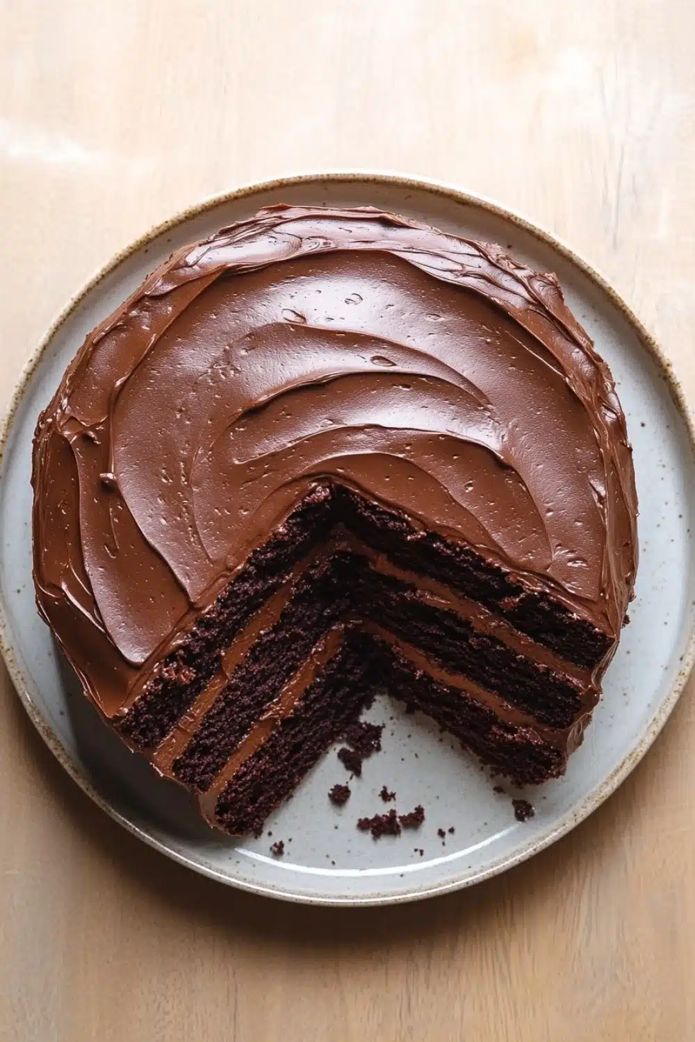 Chocolate cake normal design - the image is of a round chocolate cake on a white plate. the cake has a thick layer of chocolate frosting on top, with a swirl pattern on the sides. the frosting appears to be smooth and glossy, and the cake is arranged in a neat and orderly manner. there is a slice taken out of the cake, revealing the layers of chocolate cake. the plate is sitting on a wooden table, and there are a few crumbs scattered around the plate.