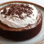 Chocolate cake tres leches - the image is of a chocolate cake on a blue and white plate. the cake is round and has a smooth texture. it is topped with a generous dollop of whipped cream and sprinkled with chocolate shavings. the plate is sitting on a white marble countertop.