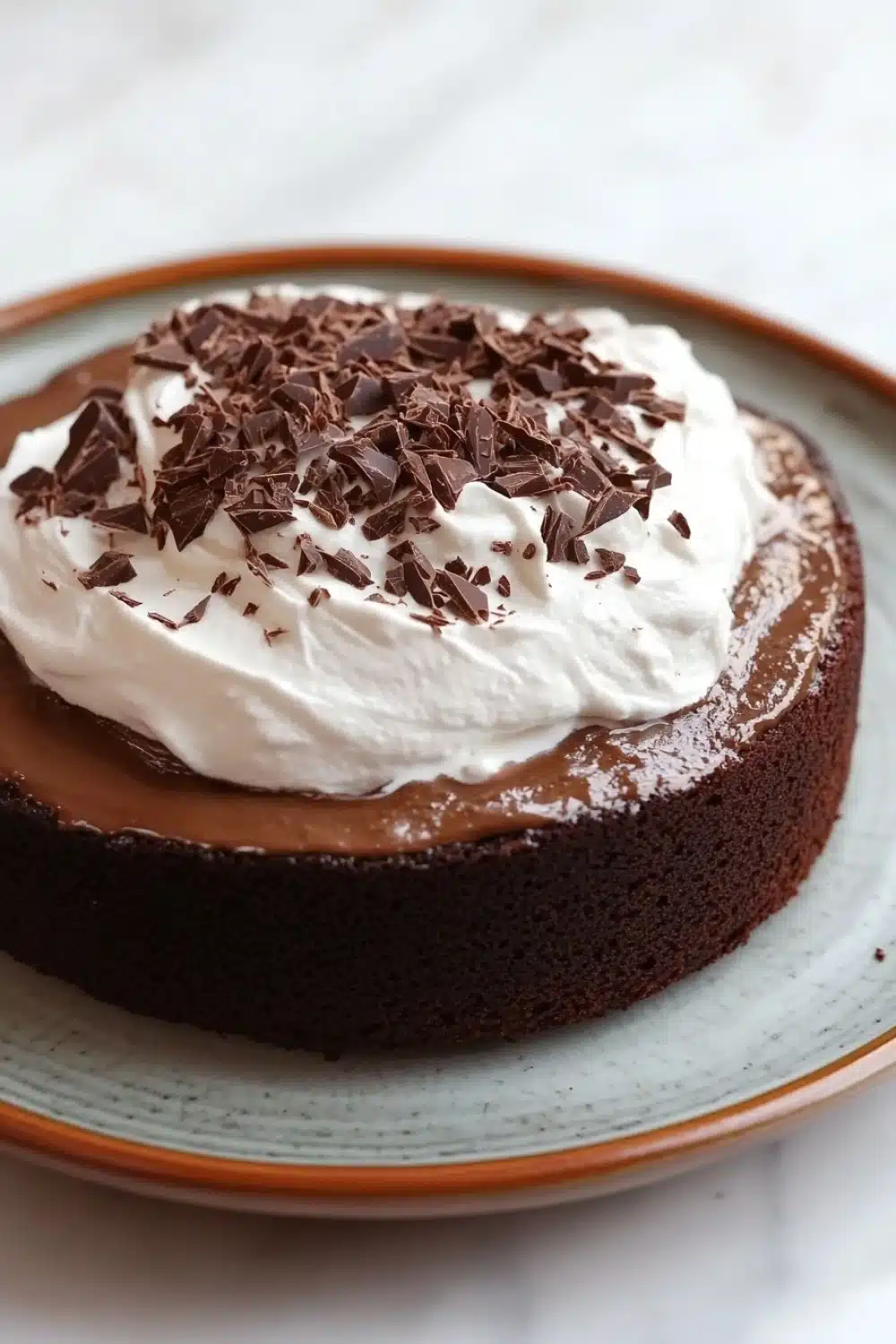 Chocolate cake tres leches - the image is of a chocolate cake on a blue and white plate. the cake is round and has a smooth texture. it is topped with a generous dollop of whipped cream and sprinkled with chocolate shavings. the plate is sitting on a white marble countertop.