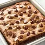 Chocolate chip cookie sheet cake - the image shows a rectangular baking tray with a freshly baked brownie in it. the brownie is golden brown in color and has a crumbly texture. on top of the brownie, there are small chocolate chips scattered across the surface. the tray is sitting on a white marble countertop.