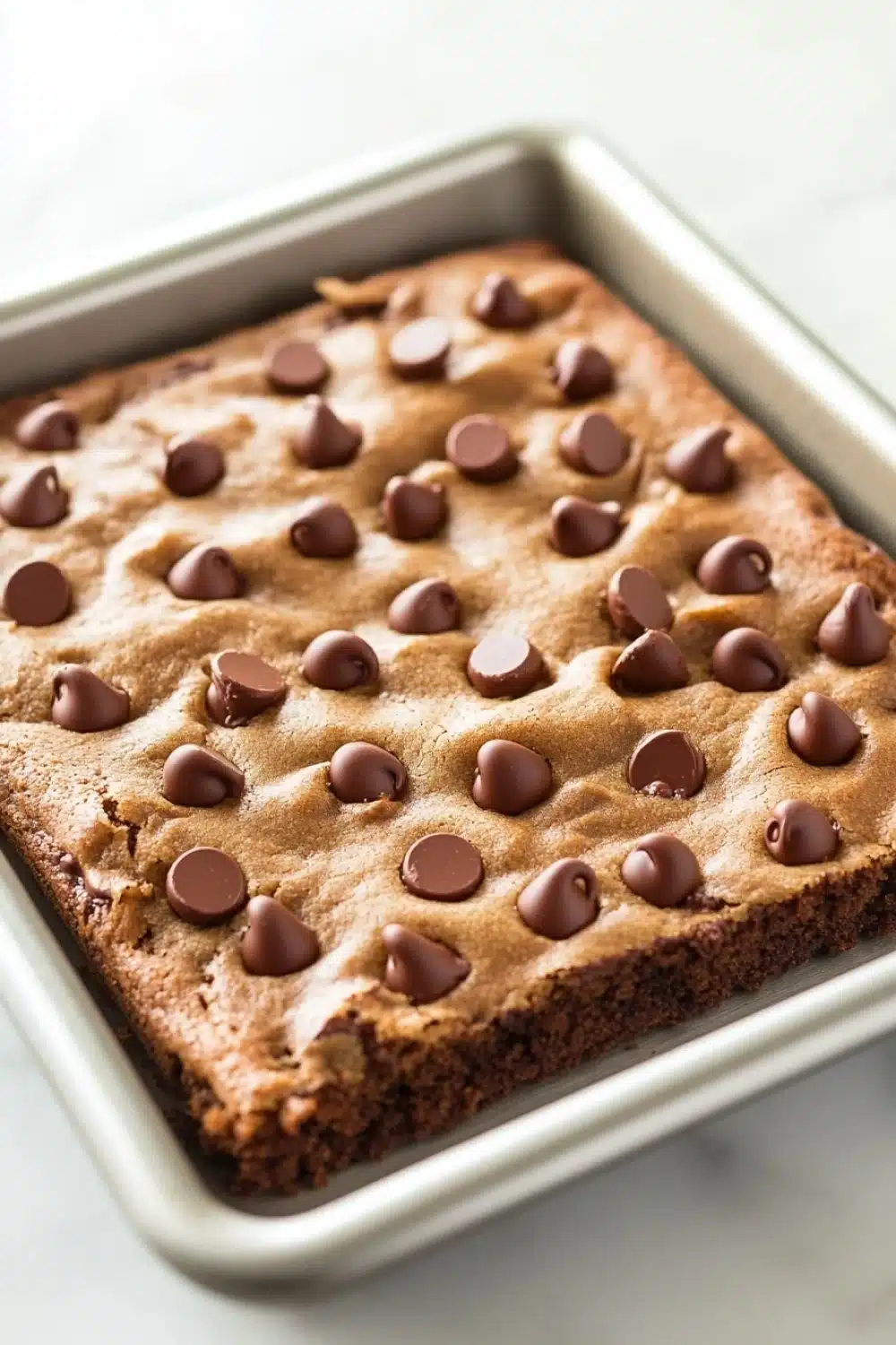 Chocolate chip cookie sheet cake - the image shows a rectangular baking tray with a freshly baked brownie in it. the brownie is golden brown in color and has a crumbly texture. on top of the brownie, there are small chocolate chips scattered across the surface. the tray is sitting on a white marble countertop.