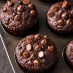 Chocolate muffin vegan - the image shows a muffin tin filled with freshly baked chocolate cupcakes. the cupcakes are round and have a golden brown color. they are topped with small chocolate chips scattered across top, giving them a crunchy texture. the tin is sitting on a wooden table, and the background is blurred, making the cupcakes the focal point of the image.
