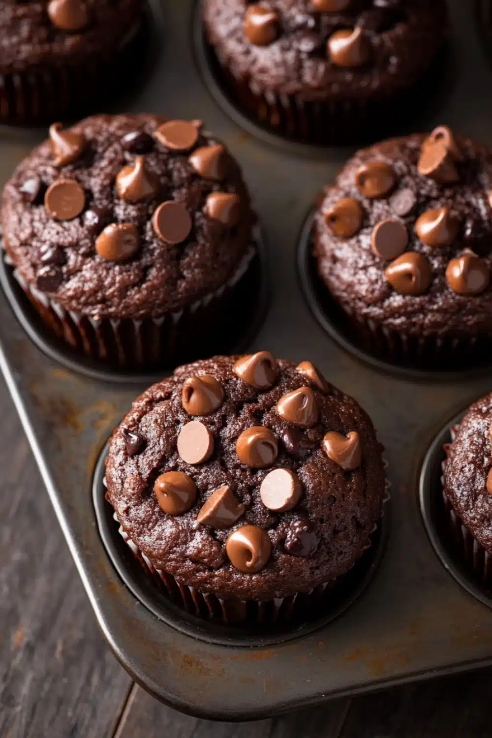 Chocolate muffin vegan - the image shows a muffin tin filled with freshly baked chocolate cupcakes. the cupcakes are round and have a golden brown color. they are topped with small chocolate chips scattered across top, giving them a crunchy texture. the tin is sitting on a wooden table, and the background is blurred, making the cupcakes the focal point of the image.