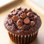 Chocolate muffin without eggs - the image is of a chocolate muffin on a gold tray. the muffin is round and has a golden brown color. it is topped with small chocolate chips scattered across its surface. the chocolate chips are arranged in a circular pattern, with some overlapping each other. the tray is a light beige color and appears to be made of parchment paper. the background is blurred, so the focus is on the muffin.