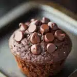 Chocolate quinoa muffin - the image is a close-up of a chocolate muffin on a baking tray. the muffin is round and has a golden brown color. it is topped with small, round chocolate chips arranged in a circular pattern. the chips are evenly spaced and appear to be freshly baked. the tray is lined with parchment paper and the background is blurred, making the muffin the focal point of the image.