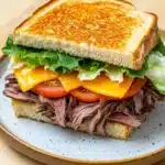 Club sandwich beef - the image shows a sandwich on a blue and white plate. the sandwich is made with two slices of toasted bread, filled with a generous portion of roast beef, lettuce, tomato, and cheese. the beef is cooked medium-rare and appears to be seasoned with herbs and spices. the lettuce is a mix of green and yellow, and the tomato is a bright red. the cheese is melted and oozing out of the sides of the sandwich. the plate is sitting on a wooden table.
