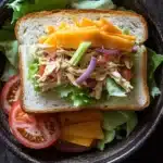 Club sandwich ensalada de pollo - the image is a close-up of a sandwich in a black bowl. the sandwich is made with two slices of white bread and is filled with a variety of ingredients. on top of the bread, there is a layer of shredded chicken, sliced carrots, red onion, and sliced tomatoes. the chicken appears to be seasoned with herbs and spices, and the vegetables are arranged in a colorful and appetizing manner. the bowl is sitting on a dark wooden table.
