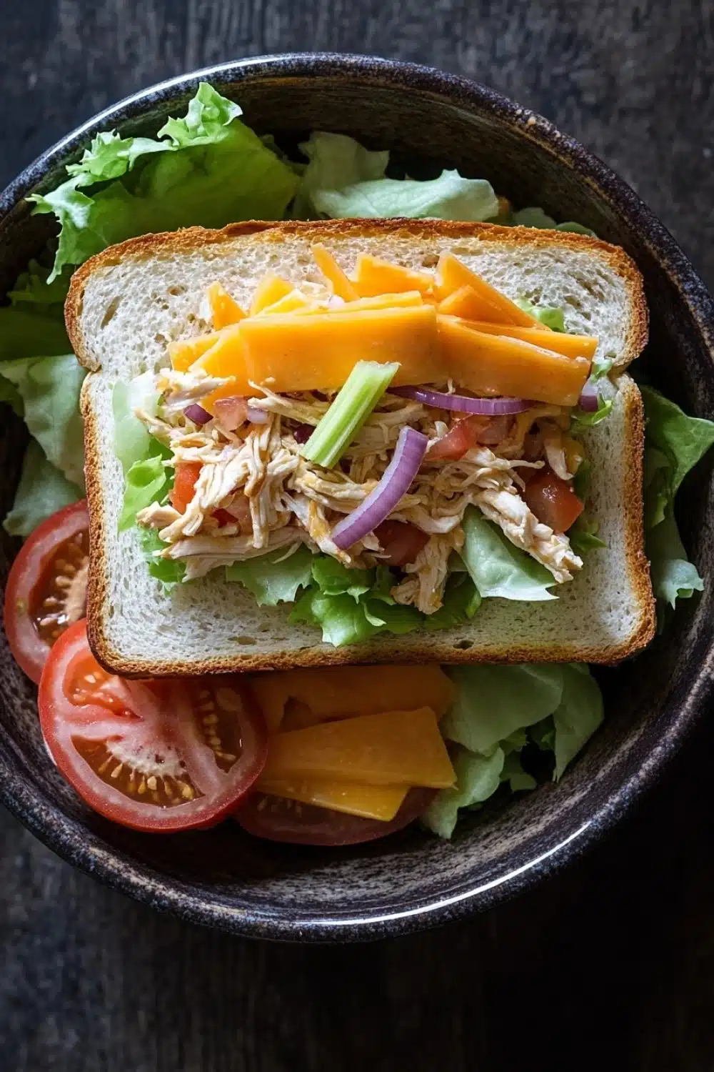 Club sandwich ensalada de pollo - the image is a close-up of a sandwich in a black bowl. the sandwich is made with two slices of white bread and is filled with a variety of ingredients. on top of the bread, there is a layer of shredded chicken, sliced carrots, red onion, and sliced tomatoes. the chicken appears to be seasoned with herbs and spices, and the vegetables are arranged in a colorful and appetizing manner. the bowl is sitting on a dark wooden table.
