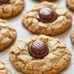 Cookie peanut butter hersheys kisses - the image shows a tray of freshly baked peanut butter cookies. the cookies are round and golden brown in color, with a crumbly texture. they are arranged in a single layer on the tray, with some overlapping each other. in the center of each cookie, there is a chocolate-covered oreo cookie with the letter "m" on it. the cookie is drizzled with a dark chocolate sauce. the tray is lined with parchment paper, and the background is a wooden table.