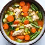 Crock pot chicken vegetable soup - the image is a close-up of a bowl of soup. the soup appears to be a vegetable soup with chunks of chicken, carrots, green beans, corn, and zucchini. the vegetables are mixed in with the soup, creating a colorful and appetizing appearance. the bowl is made of ceramic and is sitting on a white marble countertop.