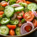 Cucumber tomato onion salad lemon - the image is a close-up of a colorful salad in a bowl. the salad is made up of sliced cucumbers, red onions, and cherry tomatoes. the cucumbers are cut into thin slices and are arranged in a circular pattern. the tomatoes are bright red and appear to be ripe and juicy. the red onions are sliced into thin strips and are scattered throughout the salad. there are also some fresh herbs, possibly parsley, adding a pop of green color to the dish. the bowl is made of dark wood and is sitting on a black countertop.