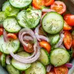 Cucumber tomato onion salad with ranch - the image is a close-up of a colorful salad in a bowl. the salad is made up of sliced cucumbers, red onions, and cherry tomatoes. the cucumbers are arranged in a circular pattern, with some overlapping each other. the red onions are sliced into thin strips and are scattered throughout the salad. the cherry tomatoes are bright red and appear to be ripe and juicy. there are also some dill seeds sprinkled on top, adding a pop of green color to the dish. the bowl is sitting on a white tablecloth with a blue and white striped napkin underneath.