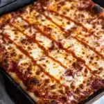 Detroit style pizza in cast iron - the image shows a rectangular baking dish with a freshly baked pizza in it. the pizza has a golden brown crust and is covered in melted cheese and tomato sauce. the cheese is melted and bubbly, and the tomato sauce is drizzled over the top. the dish is sitting on a white marble countertop with a gray cloth napkin on the side. the background is blurred, but it appears to be a kitchen countertop.