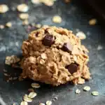 Edible cookie dough with oatmeal - the image shows a single cookie on a black baking tray. the cookie appears to be freshly baked and has a golden brown color. it is topped with chocolate chips and oats, giving it a crunchy texture. the tray is lined with parchment paper, and there are scattered oats around the cookie. the background is dark, making the cookie stand out.